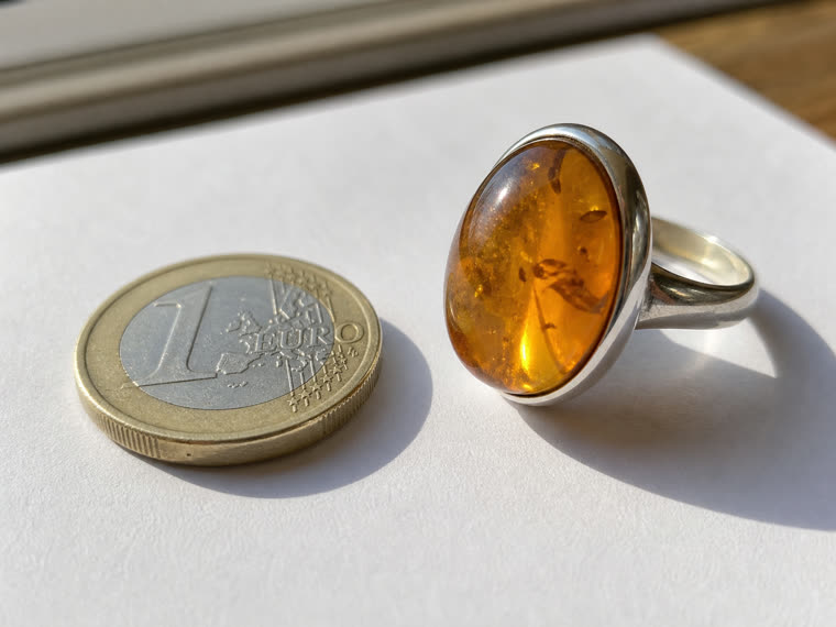 1-Euro-Münze neben einem Bernstein-Cabochon-Ring in Silberfassung als Größen-Referenz, ausgelegt auf weißem Papier am Fensterlicht