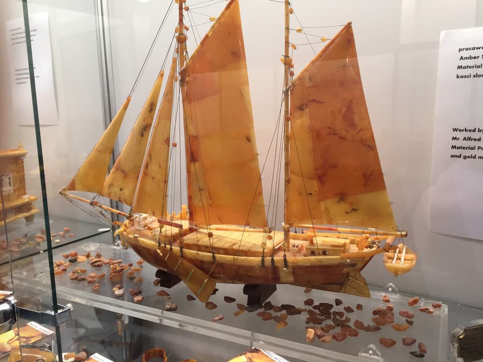 Detailliertes Bernstein-Schiffsmodell mit Bernstein-Segeln und Roh-Bernstein-Splittern als symbolische Wellen, ausgestellt in einer Museumsvitrine, Sinnbild für die maritime Bernsteinhandels-Tradition