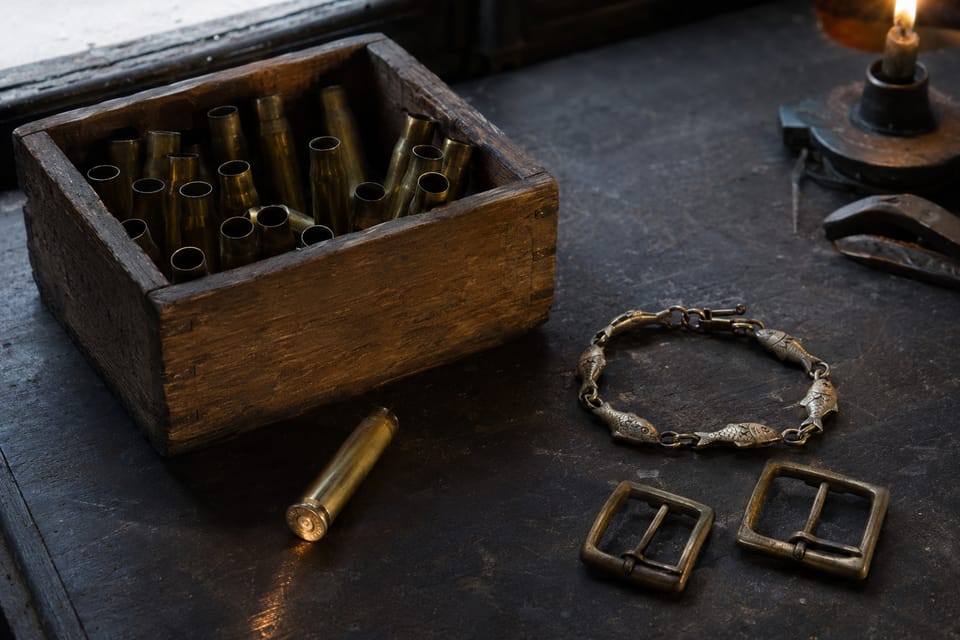 Post-war still life, 1946: a wooden crate of empty brass shell casings, beside it a link bracelet with fish motifs and two brass belt buckles by candlelight