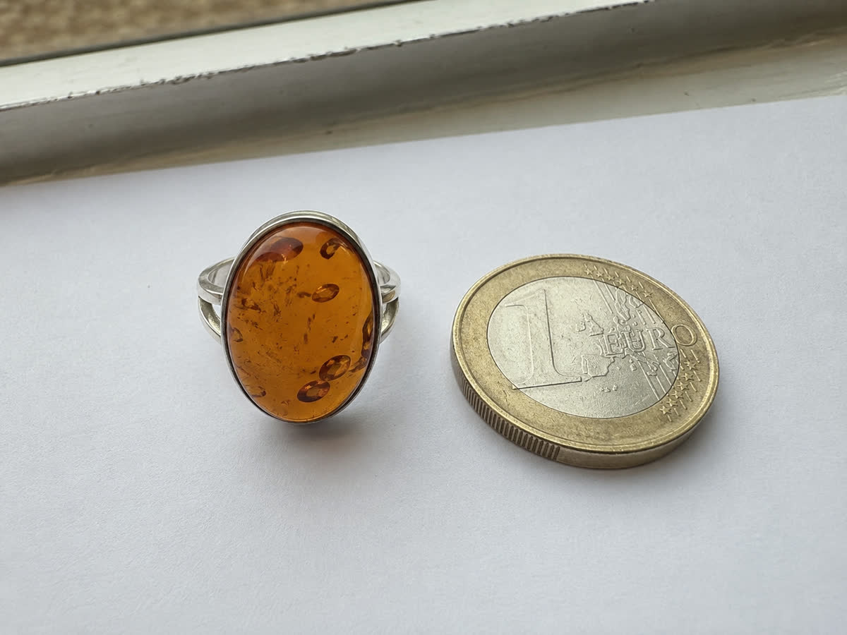Honig-Cognac Bernstein-Cabochon in Silberring-Fassung neben einer 1-Euro-Münze als Größen-Referenz auf weißem Papier am Fensterlicht