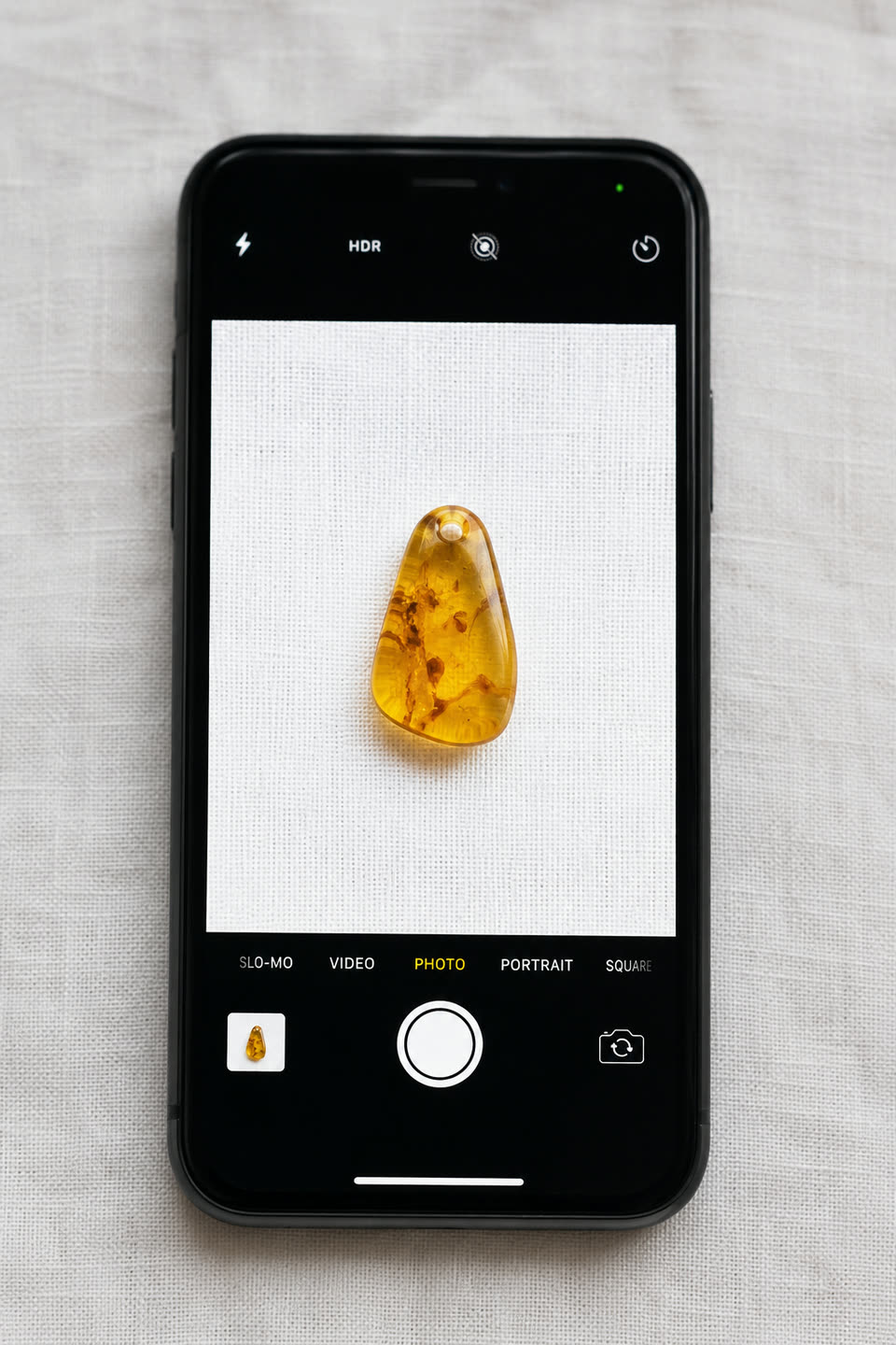 iPhone-Kamera-Viewfinder zeigt einen butterscotch-honigfarbenen baltischen Bernstein zentriert auf hellem Leinen-Hintergrund, Demonstration der korrekten Aufnahme für die Foto-Bewertung: neutraler Hintergrund, mittig platziert, gutes Tageslicht
