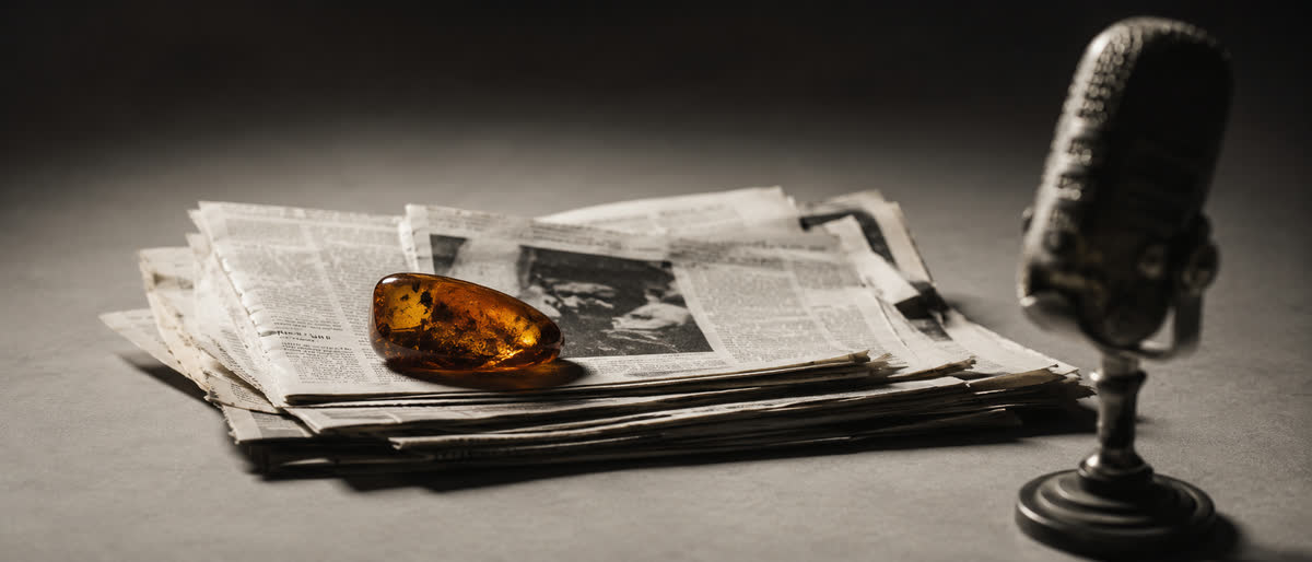 Polierter baltischer Bernstein als Briefbeschwerer auf einem Stapel deutscher Zeitungsausschnitte, daneben ein Vintage-Studio-Mikrofon im Halbschatten