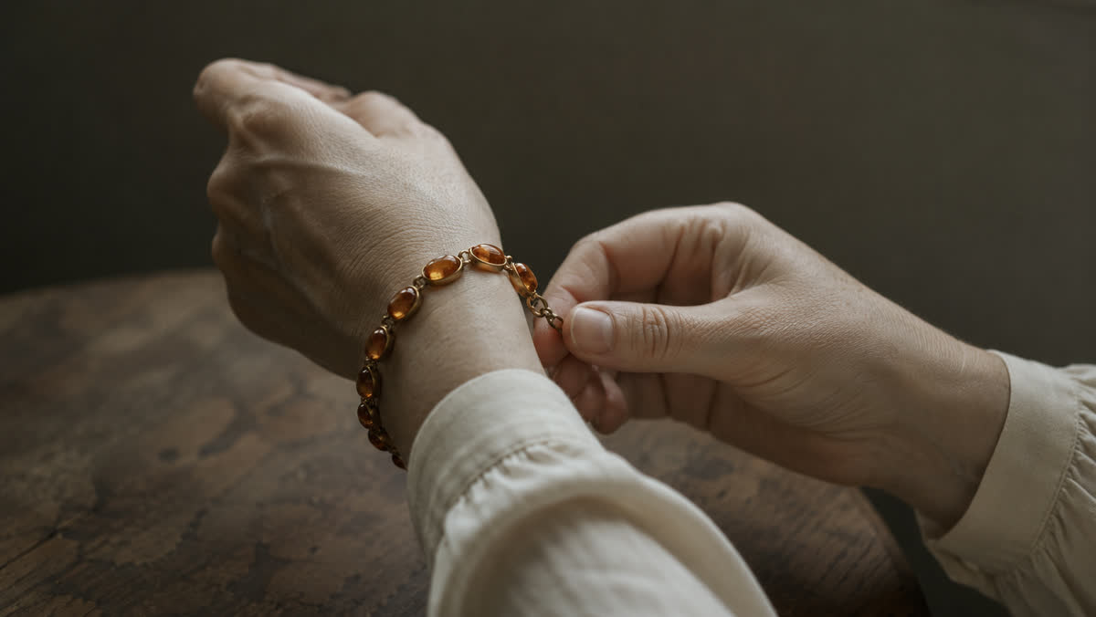 Zwei Hände beim Anlegen eines vintage Bernstein-Cabochon-Armbands am linken Handgelenk, nur Hände und Unterarme sichtbar in cremefarbener Bluse, warmes Tageslicht, alte Holztischkante leicht unscharf im Hintergrund
