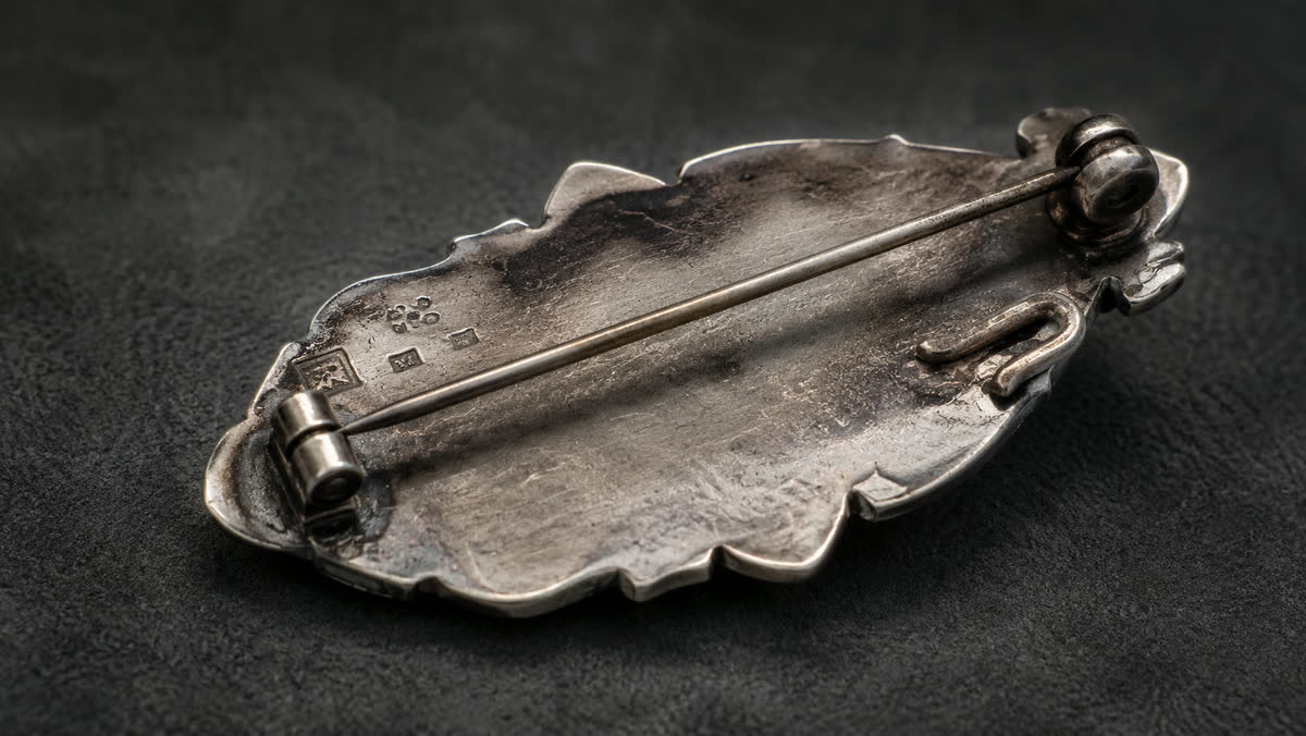 Makro der Rückseite einer antiken Bernstein-Brosche: lange Silbernadel, klassischer rollender Trommel-Verschluss, kleine Silber-Punzen am Scharnier, angelötete Sicherungs-Schlaufe, Patina aus Jahrzehnten