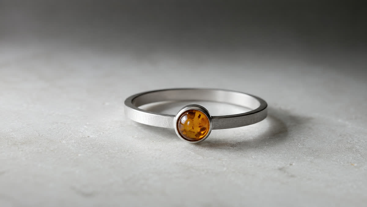 Zeitgenössischer Minimal-Ring mit schmalem gebürstetem Silberband und einem kleinen flächenbündig gefassten Honig-Bernstein-Cabochon, auf hellem Stein